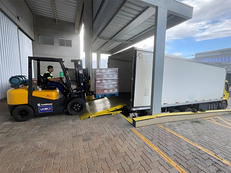 Empilhadeira realiza carregamento de carga em caminhão baú ao lado de doca, integrando etapas de distribuição logística.